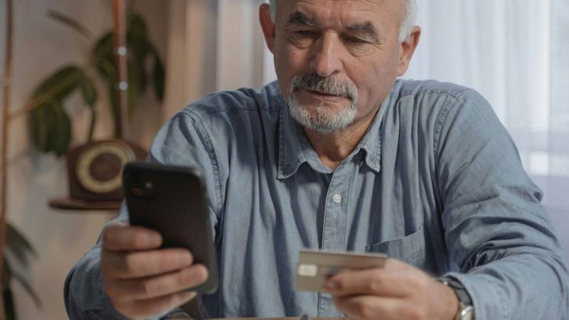 An older man using a smartphone while looking at a bank card.